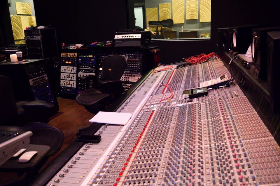 A large analogue recording console in the Los Rios control room, racks of outboard gear behind it and a live room visible through the glass