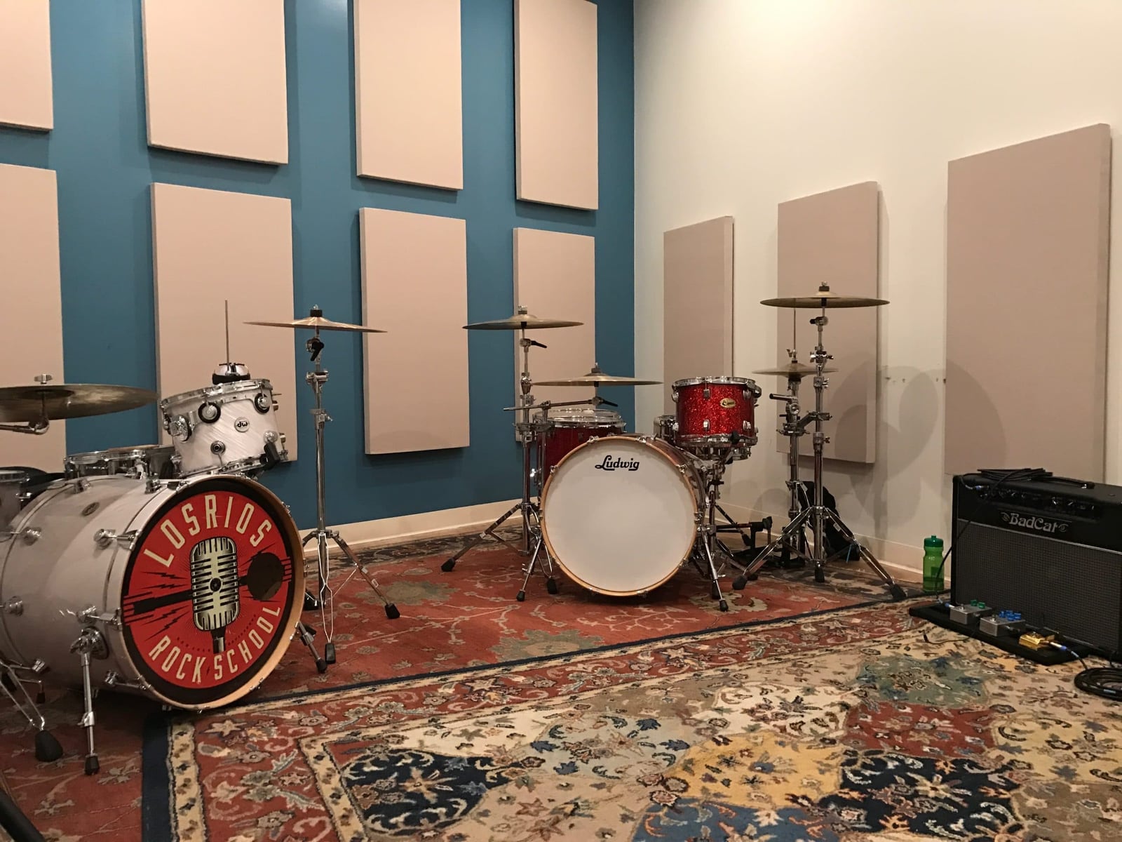 Three drum kits on a Persian rug in a Los Rios lab, the nearest bearing the Los Rios Rock School logo on its head, with a BadCat amplifier against the wall