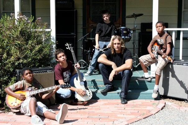 Tyler Marolf sitting on the front steps of the white clapboard Los Rios cottage with five young students holding guitars and a bass, a drum kit set up on the porch behind them