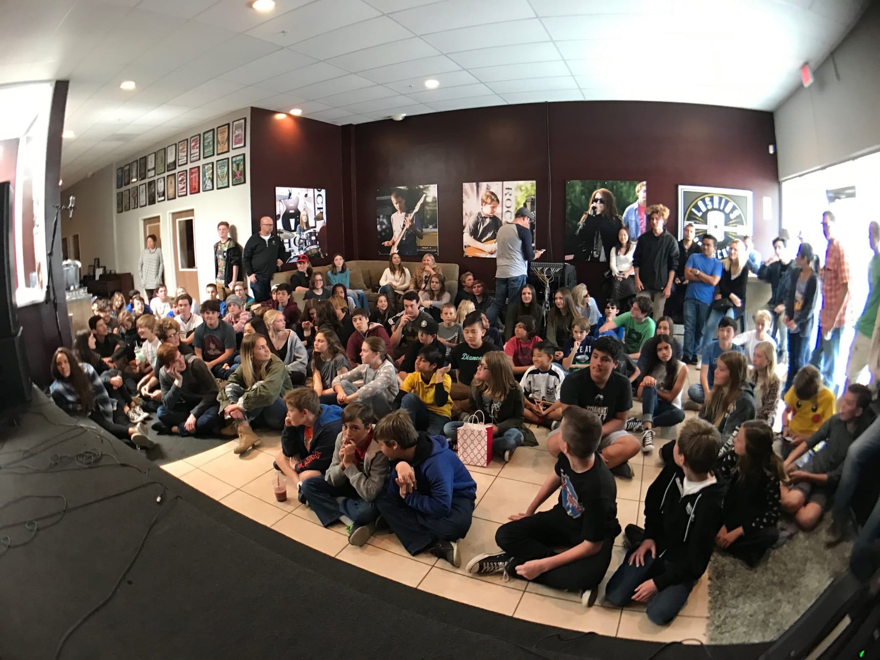 A hundred students sitting on the floor of the Los Rios Rock School stage room watching a performance, with framed concert posters hung in a row along the wall behind them